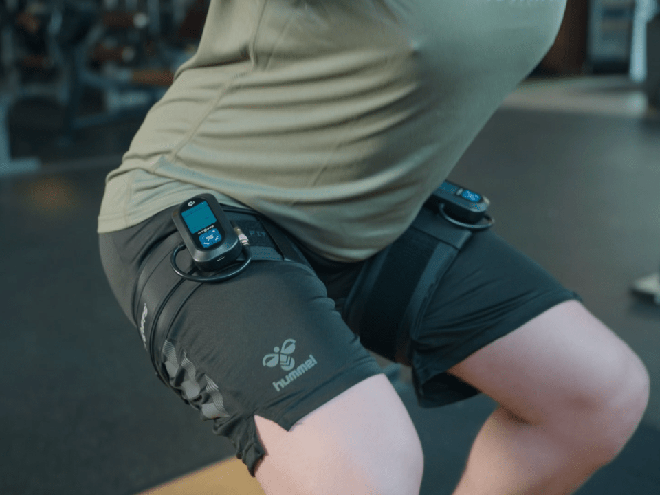 Person performing a squat exercise in a gym while using BFR Unit and Leg Cuffs V4.0 on their thighs, equipped with electronic devices monitoring blood flow restriction during training