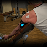 Person performing shoulder rehabilitation exercises using a preacher curl bench while wearing a BFR (Blood Flow Restriction) unit with Arm Cuff V4.0 and electrode patches attached to the shoulder