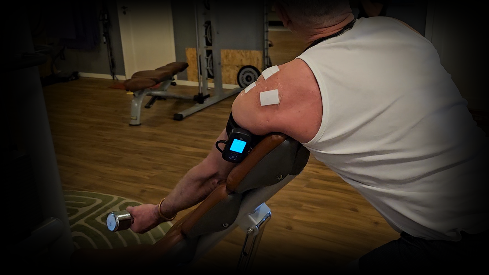 Person performing shoulder rehabilitation exercises using a preacher curl bench while wearing a BFR (Blood Flow Restriction) unit with Arm Cuff V4.0 and electrode patches attached to the shoulder