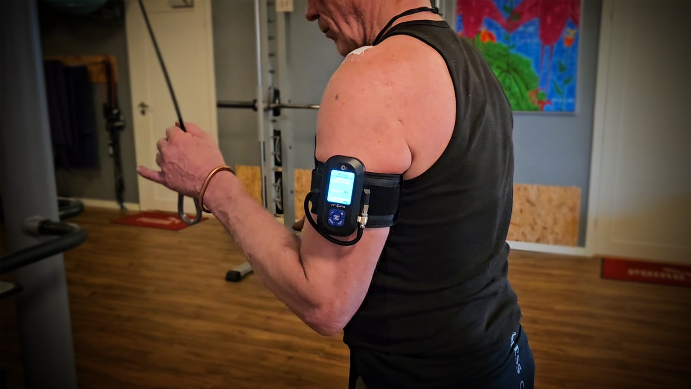 Man performing triceps cable extensions in a gym wearing a BFR (Blood Flow Restriction) Unit with Arm Cuff V4.0. The device is being used as part of rehabilitation therapy following subacromial decompression surgery. The BFR unit is secured to the upper arm, with a digital display visible, helping monitor and control the therapy session